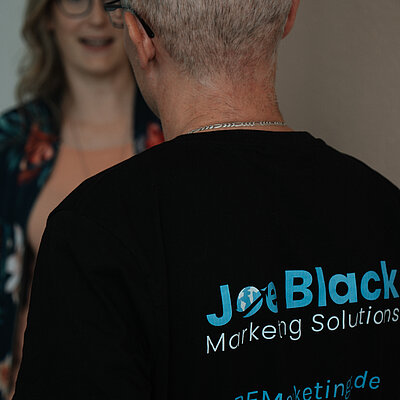 Joe Black und Helen Bauer im Gespräch. Die Rückenaufschrift des Shirts von Joe Black im Fokus mit Infos zu seinem Unternehmen