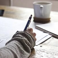 Sinnbild für Schreiben mit der Hand, Lernen und Hausaufgaben. Gezeigt wird eine Hand, die einen Stift hält. Papier liegt auf einem Schreibtisch. Dort steht auch eine Tasse.