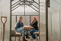 Nestbau-Koordinatorin Helen Bauer und Zuzüglerin Becky Hellwig im Gespräch. Dabei sitzen sie in einem Gewächshaus, welches als Sitzecke mit Sand unter den Füßen umfunktioniert wurde.