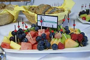 Foto zeigt Detail eines Frühstücksbuffets. Es handelt sich dabei um einen Teller mit Obstspießen und einem Hinweisschild auf den Partyservice Friedemann. Zu sehen sind Melone, Weintrauben und Erdbeeren.