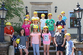 Foto zeigt eine Gruppe von Kindern auf einer großen Treppe. Die Mädchen und Jugen tragen gelbe Bauhelme aus Plastik. 