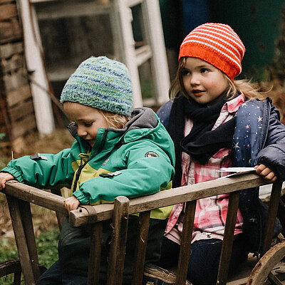 Die Kinder Leyma und Jul sitzen im Holzwagen und lassen sich durch den Garten fahren.