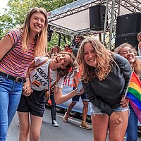 Foto zeigt eine Gruppe junger lachender Frauen auf einer Straße. Sie tragen eine Fahne mit Regenbogenfarben bei sich.