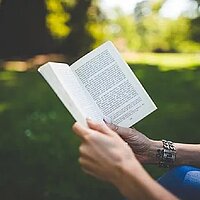 Beschäftigung mit Literatur: Foto zeigt Hände und Arme, die ein Buch halten. Im Hintergurnd ist eine Parklandschaft zu erahnen. Viel Grün mit Tiefenunschärfe.