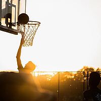 Foto zeigt Ballsport, abgebildet ist ein Basketballkorb und ein Spieler im Gegenlicht bei Sonnenuntergang.