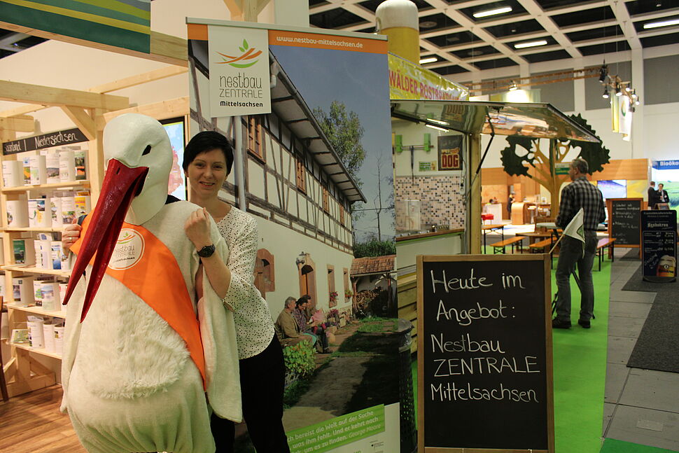 Foto zeigt Projektkoordinatporin Josefine Tzschoppe, welche die Nestbau-Zentrale Mittelsachsen auf der Internationalen Grünen Woche in Berlin präsentiert. Am Informationsstand wird die junge Frau von einer Person in einem Storchenkostüm unterstützt.