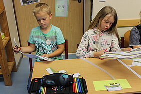 Kinderbeschäftigung in einer Grundschule: Foto zeigt einen Jungen und ein Mädchen mit Bastelvorlagen für ein Haus. Projekttag an einer Grundschule.