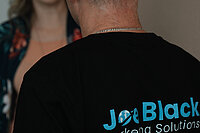 Joe Black und Helen Bauer im Gespräch. Die Rückenaufschrift des Shirts von Joe Black im Fokus mit Infos zu seinem Unternehmen