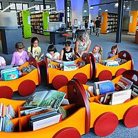 Foto zeigt das Innere einer Bibliothek, in der Kinder einer Gruppe von einer Frau zum Lesen angeleitet werden